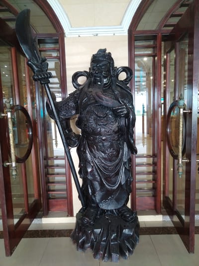 A statue of a Chinese warrior stands in the entrance of a building in Chu Văn An Ward, Vietnam. The statue is black, carved with intricate details, and stands on a rocky base. The warrior has a long beard and wears elaborate armor. He holds a large polearm with a curved blade. The building has a light-colored interior and features a set of double doors made of wood with glass panels and round handles. The doors are partially open. The floor is tiled in a light color.