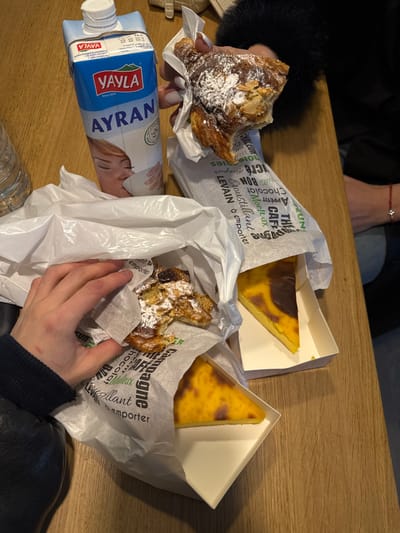 The image displays a light brown wooden tabletop laden with food and beverages, suggesting an indoor setting in Asnières-sur-Seine, France. Two individuals are present, evidenced by visible hands and lower arms. On the left, a hand with a dark sleeve is reaching into a white plastic bag containing a partially consumed almond croissant, dusted with powdered sugar and wrapped in white paper printed with French bakery terms such as "BONJOUR" and "LEVAI N à emporter". Adjacent is a partially visible clear, ribbed plastic bottle. Centrally located is a full carton of "Yayla Ayran" yogurt drink with a white screw cap, featuring an image of a person drinking. To the right, a second hand with light-colored manicured fingernails and a dark, possibly furry, sleeve holds another partially eaten almond croissant, similarly powdered. A red string bracelet is visible on this person's wrist. Also visible are two triangular slices of flan pâtissier (custard tart) in white cardboard containers, one fully exposed, the other partially within a white plastic bag, both displaying a caramelized top surface. The specific establishment within Asnières-sur-Seine is not identifiable from the image content.