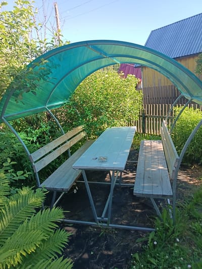 The image depicts a picnic table and benches situated outdoors under a green, arched canopy. The setting appears to be a rural or suburban area, possibly a backyard or garden in Koryazhemka, Russia, given the context.

**Objects:**
*   **Picnic Table:** A rectangular table with a white or light-colored surface, supported by a grey metal frame.
*   **Benches:** Two wooden benches, one on each side of the table, also supported by grey metal frames.
*   **Canopy:** A curved canopy made of green translucent material, supported by grey metal arches.
*   **Vegetation:** Lush green foliage, including trees and bushes, surrounds the picnic area. There are also ferns visible in the foreground.
*   **Structures:** In the background, there are parts of residential buildings with corrugated metal roofs and wooden fences. One building has a yellow-painted wooden wall.
*   **Other items on the table:** A small ashtray containing cigarette butts, and a matchbox.

**Environment:**
The scene is outdoors during daylight, under a clear blue sky. The sun casts shadows, indicating a bright day. The ground beneath the canopy appears to be covered with dark material, possibly a tarp or weed barrier, with some loose soil and scattered vegetation. The surrounding environment is characterized by abundant greenery, suggesting a well-established garden or natural growth.

**Actions/Interactions:**
No people are present in the image. The scene depicts a static setup for outdoor leisure, with objects arranged for use at a picnic table. The presence of the ashtray and matchbox suggests recent or potential use for smoking.