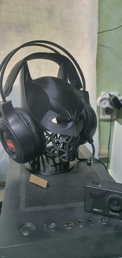 A black headset is placed on top of a black Batman-themed headphone stand. To the right of the headphone stand, there is a black action camera with "hoco." branding. A small, rectangular USB flash drive is located on the top surface of a black computer tower in the foreground. The computer tower has several USB ports and buttons visible. In the background, there is a white electrical outlet with a black plug adapter inserted. The scene appears to be an indoor setting, possibly a desk or workspace, and it is not possible to determine the specific location within Andijan City, Uzbekistan, from the image alone.