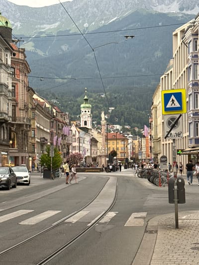 This is a vertical, eye-level, medium-wide shot of a European city street with mountains in the background. The image is captured in daylight, with overcast skies suggesting a neutral or cool weather.

The street is paved with cobblestones on the right side and asphalt on the left, with tram tracks running down the center. Several people are visible on the street. Two individuals are crossing a pedestrian crossing, one wearing a light-colored top and dark shorts, and the other in a yellow top and blue shorts. Other pedestrians are walking along the sidewalks, and a line of bicycles is parked on the right. Cars are also present on the street, including a white van and a dark sedan.

The buildings lining the street are tall and multi-story, with a mix of architectural styles, some appearing older with ornate details and others more modern. One building features a clock tower with a green dome. Flags in various colors, including pink and purple, are hung from some of the buildings.

In the background, a lush green mountain range dominates the view, partially obscured by buildings in the mid-ground. The scene appears to be in a bustling urban area with a clear view of the natural landscape.

There is a yellow diamond-shaped traffic sign with a blue border displaying a pedestrian symbol. Another sign nearby has a circular symbol with a diagonal line through it, and the word "Zone" is visible on a separate sign. The overall atmosphere of the image is that of a lively city with a strong connection to its natural surroundings.
