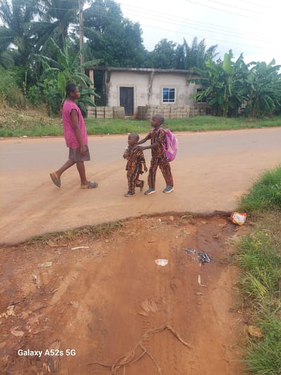 Three children are walking on a dirt road in Umudioka, Nigeria. The child on the left is wearing a pink and black striped shirt and dark shorts. The child in the middle is wearing a colorful patterned outfit and carrying a pink backpack. The child on the right is wearing a similar colorful patterned outfit. In the background, there is a partially constructed building with windows and a doorway. Banana trees and other foliage surround the area. The road appears to be made of reddish-brown dirt, with some grass and debris visible along the edges. A single power line extends across the top of the image.