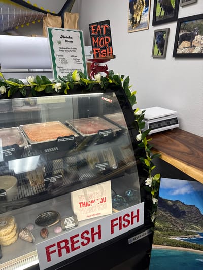 This image depicts the interior of a commercial food establishment, likely a deli or fish market, located in Port Allen, Kauai, Hawaii, USA. The primary subject is a refrigerated display case in the foreground, adorned with artificial green foliage and white flowers along its top edge.
Inside the glass display case, multiple food items are visible across three shelves. The top shelf contains several rectangular metal trays covered with clear plastic wrap, identified by labels as "CHILI GARLIC POKE," "SPICY MAYO POKE," and "HAWAIIAN STYLE POKE." The middle shelf holds pre-packaged items, including "TURKEY W/ HAVARTI $6.95" and "HAM W/ AMERICAN CHEESES $6.95," alongside a white container labeled "SOUP." The bottom shelf displays a variety of items: stacks of circular food items (possibly cookies), a black abalone shell, two conch shells, three pink scallop shells, a small white sign indicating "REUSABLE COOLER BAG OR ICE $3.50," and a white paper bag printed with "THANK YOU THANK YOU THANK YOU Have a Nice Day" in red text.
Above the display case, a white sign with a green border advertises "Matcha Latte" with pricing and flavor options. Adjacent to it, a black sign on a red crab-shaped stand reads "EAT MOR FISH" in red lettering. Partially visible above the Matcha Latte sign is a mounted yellow and blue fish decoration, likely a marlin or tuna. Two brown paper bags are positioned behind the Matcha Latte sign.
The background wall is white and features a collection of framed photographs depicting various animals, including several dark-furred canines and a wild boar. On a dark wooden countertop to the right, a white digital scale is present. A large print or poster of a scenic Hawaiian coastline with green mountains, a sandy beach, and blue water leans against the right side of the display case. A prominent red sign reading "FRESH FISH" in block letters is attached to the lower front of the display case, with "BEVERAGE-AIR" visible below it. No individuals are present in the scene.