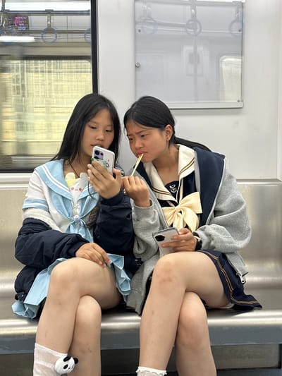 Two young Asian girls in school uniforms are sitting next to each other on a subway train. One girl is holding chopsticks and the other a mobile phone. The background shows the interior of a subway car. The location is Tiaodeng, China.