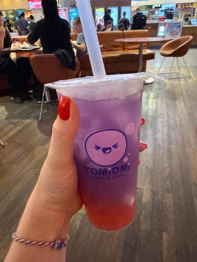 The image shows a person holding a Tomiomi Bubble Cafe drink in what appears to be a cafe setting. The drink is layered, with a purple-colored layer at the top and a red-colored layer at the bottom. The person has red nail polish and is wearing a bracelet with red, white, and blue braided string. The cafe setting shows other patrons and furniture in the background. The location is implied by the cafe's name to be in Pavlovskaya, Russia.