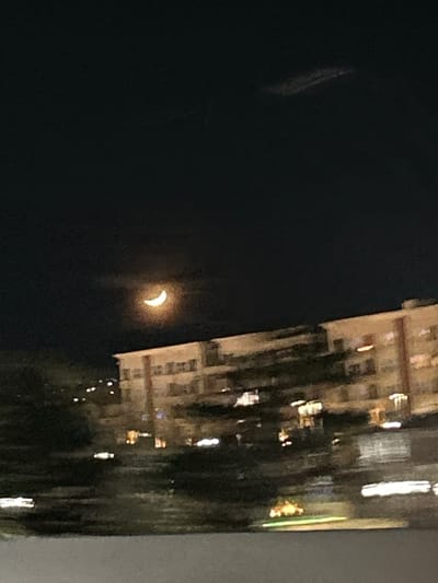 The image displays a nighttime scene within the city of Sparks, USA. A bright, yellowish crescent moon is centrally positioned in the upper-left portion of a dark sky. A faint, elongated cloud formation is discernible in the upper-right sky. In the mid-ground, several multi-story buildings, consistent with residential or commercial structures, span horizontally from the mid-left to the right edge of the frame. These buildings feature numerous illuminated windows and exhibit significant horizontal motion blur. Dark, indistinct masses resembling vegetation are present in the foreground and beneath the building structures. Additional scattered artificial lights are visible on the distant left horizon. The bottom edge of the image features an unidentifiable light-colored horizontal surface. No individuals, specific actions, or direct interactions are discernible within the frame. The overall image quality, characterized by motion blur and low light, suggests capture from a moving platform.