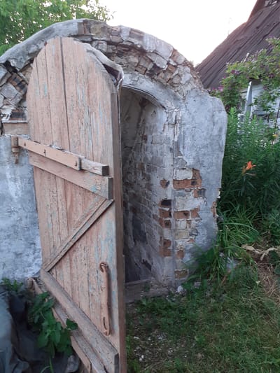 The image shows the weathered exterior of an old, possibly disused, structure in Deremezna, Ukraine. The most prominent feature is a large, arched doorway, partially obscured by a set of wooden double doors. These doors are painted a faded pinkish-orange, with visible signs of peeling paint and age. They are reinforced with diagonal wooden planks, forming a triangle on the right-hand door, and secured with simple metal clasps.

The archway itself appears to be constructed of brick and stone, with sections of crumbling plaster revealing the underlying red bricks. The arch's surface is rough and irregular, indicating significant wear and tear. Greenery, including grass and weeds, grows around the base of the structure and along the top of the archway, suggesting it is in a rural or overgrown setting. In the background, more dense vegetation can be seen, with hints of purple flowers and a single orange flower. The lighting suggests it might be late afternoon or early evening, with a soft, natural light illuminating the scene. There is no visible text in the image. The overall impression is one of neglect and the passage of time.