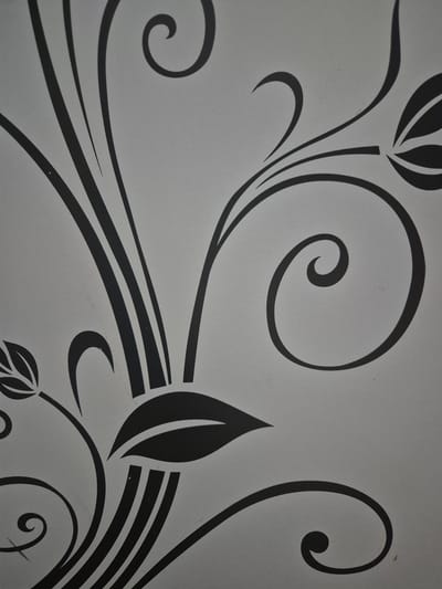 A black floral stencil is applied to a gray surface. The design features swirling vines, stylized leaves, and thin, curving lines. The stencil is abstract and decorative, with no discernible real-world context such as a specific location in Hromtau, Kazakhstan. The image is a close-up of the design, showing its texture and detail. There are no people or actions depicted.