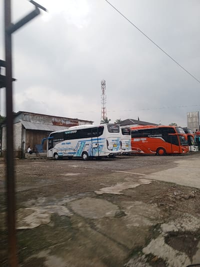 The scene depicts an outdoor parking area, stated to be in the city of Qorday, Kazakhstan, under an overcast sky. In the immediate foreground, a dark, vertical metal structure partially obstructs the left side of the frame. The ground comprises compacted dirt, gravel, and irregular sections of cracked asphalt or concrete.
The midground features a line of multiple large passenger buses. The two most prominent buses on the left are predominantly white with blue and black graphic accents, bearing the branding "DENY TRANS" and "HIGH DECK". To their right and further back are several orange buses, also visibly branded "DENY TRANS". To the far left, behind the front-most white bus, is a single-story building constructed with corrugated metal siding and a matching roof. Two individuals are seated on a low ledge or step directly outside this building; their specific actions are not discernible.
In the background, a tall, red and white lattice-style telecommunications tower stands centrally. Patches of green vegetation are visible behind the tower and other structures. Another building with a dark roofline is partially obscured behind the orange buses.