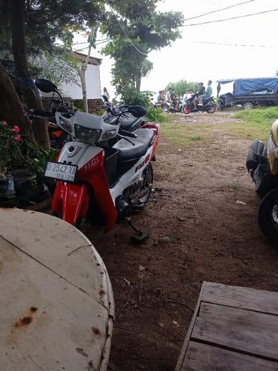 The image shows a white and red motorcycle with the license plate number D 2542 YA 05-29 parked on a dirt road. There are other motorcycles parked nearby, and several people can be seen in the distance, seemingly riding or walking away.  The image was taken in Pameungpeuk, Indonesia, at coordinates -7.668, 107.694. The ground is a light brown color and the area is partially covered in vegetation. There is a large wooden platform on the right side of the image, and a round wooden table with a faded white surface in the foreground.