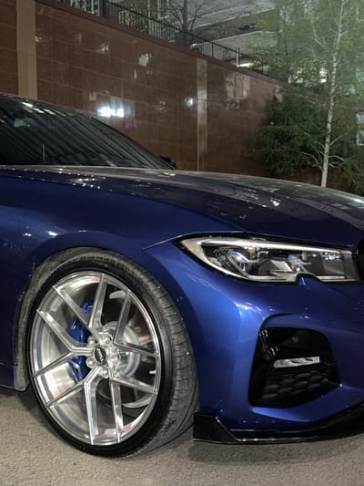 The image shows a close-up of the front of a blue car, focusing on the headlight, front fender, and wheel. The wheel has a silver multi-spoke design and blue brake calipers. The car is parked on what appears to be an asphalt surface. A building with a tile facade and a tree are visible in the background. There's nothing in the image to confirm this is in Novoaleksandrovka, Russia.