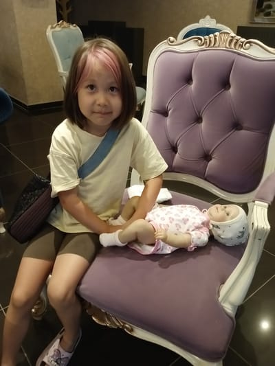 In Kırıkkale, Turkey, a young girl with light brown hair with pink highlights sits on a plush purple armchair. She is wearing a light yellow t-shirt and dark shorts, with a blue strap over her shoulder and a dark patterned bag slung across her back. To her right, a doll lies on the armchair, dressed in a pink and white patterned outfit and a white cap. The girl is looking towards the camera with a neutral expression. The armchair has ornate white and gold trim. The background is indistinct, with hints of other furniture and a tiled floor.