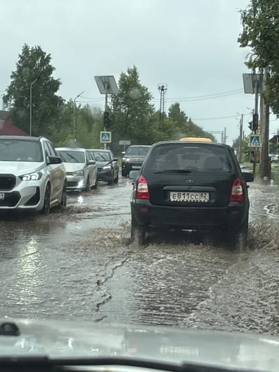 This is a photograph taken from inside a vehicle, looking forward at a street scene in Krasnokamsk, Russia. The road is heavily flooded with murky water, creating significant splashes as vehicles navigate through it. In the foreground, the hood of the vehicle from which the photo is taken is visible. Several cars are driving in a traffic jam on the flooded street. A silver SUV is in the left lane, followed by a lighter colored sedan, and then a black hatchback. The black hatchback has the license plate "E811CC 82" and appears to be a Lada. Further down the road, other vehicles are visible in the distance. Traffic lights and street signs are present on poles to the right of the road. Trees and buildings line the sides of the street. The sky is overcast and the overall atmosphere suggests a rainy day.