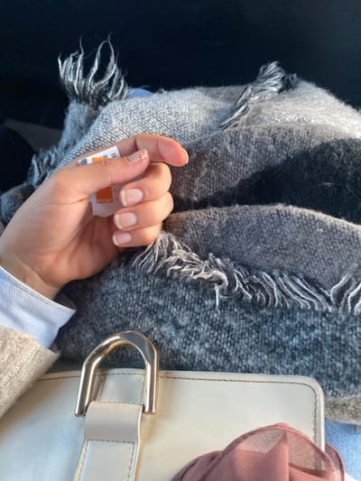 The scene depicts a close-up, downward-angled view inside what appears to be a vehicle, likely within the city of Monastir, Tunisia. A light-skinned human hand, possibly female, with unpolished fingernails showing natural white tips, is centrally positioned. The hand holds a small, torn piece of white paper displaying the word "orange" in orange text and the number "1." The hand rests upon a textured, grey-toned woven scarf or blanket with frayed, fringed edges. Below the scarf, a segment of a light cream or off-white bag with a prominent gold-colored metal D-ring or buckle is visible. To the left, a portion of a white collared shirt and a beige knitted garment are discernible, presumably worn by the individual whose hand is shown. A dark, indistinct background suggests the interior of a vehicle, with a strong shaft of sunlight illuminating the subjects from the upper right.