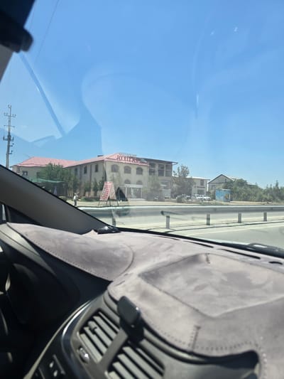 The image is taken from inside a vehicle, looking out through the windshield. The view encompasses a roadside scene with a large, light-colored building with a red roof that has signage reading "RESTAURANT JOYIZAN". A person is standing near the building, and a red A-frame sign is positioned in front of it, displaying text in a non-English script, likely Uzbek. Beyond the building, there are trees and other structures, including what appears to be a smaller building with a yellow utility box in front of it, and cars parked along the road. The road itself is visible with a metal guardrail separating it from the area with the buildings. The sky is clear and blue, indicating a sunny day. The foreground shows the interior of the car, including the dashboard with a grey, textured cover and air vents. The location is identified as Safkarda, Uzbekistan.