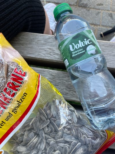 An eye-level, close-up shot shows a partially opened yellow plastic bag of sunflower seeds next to a partially filled plastic bottle of Volvic natural mineral water. The bag, labeled "Sonnenkerne," has red and white stripes along its bottom edge. The bottle has a green cap and features a label with a green soccer field design and the text "Volvic," "Natürliches Mineralwasser," and "Jetzt Gewinnen." A person's legs, clad in black textured fabric, are visible in the upper left corner, sitting on a wooden bench. The bench appears to be in an outdoor urban setting, possibly a park or public area in Waldbröl, Germany, indicated by the textured pavement and shadows suggesting daylight.