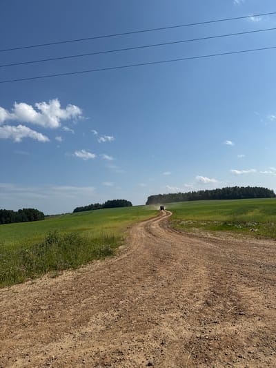 The image depicts a dirt road winding through a grassy field on a sunny day. The sky is a clear blue with scattered white clouds. Power lines are visible on the left side of the image. A dark, agricultural vehicle is driving on the dirt road in the distance. The field is bordered by a dense line of trees. The scene appears to be rural, possibly in Бабка, Russia, given the context. The time of day appears to be daytime, and the weather is sunny and fair. There is no visible text in the image.