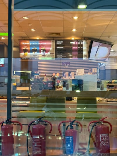 The image shows the interior of a VIVA Café, as indicated by the prominent "VIVA Café" branding on a menu board and various promotional materials. The menu board displays a selection of coffee drinks with prices in Bulgarian Leva and Euros. The café appears to be well-lit with overhead fluorescent lighting. Behind the counter, there are various food and drink items displayed, along with coffee machines and other equipment. In the foreground, three red fire extinguishers are visible, positioned in front of a glass partition. The overall environment suggests a modern, clean, and well-maintained establishment. There is no visible indication that this establishment is located in Kalugerovo, Bulgaria.