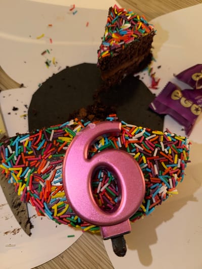 A close-up, overhead shot captures a slice of chocolate cake adorned with colorful sprinkles and a pink, metallic number "9" candle. The cake is partially placed on a white plate or box, with crumbs and loose sprinkles scattered around. In the background, portions of purple candy wrappers with gold detailing are visible. The scene appears to be indoors, possibly in a home, as suggested by the wooden surface visible at the bottom right. The lighting is warm, casting a shadow of the candle and cake onto the surface, indicating it might be daytime or artificial lighting. The overall impression is a celebratory dessert, likely for a birthday, with the number "9" clearly indicating a 9th birthday. The context provided places the image in Ashdod, Israel.