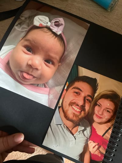The image displays an open, coil-bound black photo album or scrapbook, viewed from an oblique angle. The album contains two glossy photographs. The upper photograph features a close-up of an infant with dark hair, wearing a pink headband adorned with a multi-patterned bow. The infant's eyes are open, and its tongue is extended slightly from its mouth. The lower photograph is a selfie-style portrait of two adults: a man and a woman. The man has a beard, dark hair, a lower lip piercing, and is smiling, wearing a light-colored polo shirt. The woman has blonde hair, a nose piercing, a white substance on her left cheek and chin, and is smiling while wearing a red top. Her left hand is visible, seemingly interacting with the man's arm. A human hand, with a visible thumb and fingers, is holding the lower-left edge of the album. The background behind the album appears to be a light-colored wooden surface, with a partial blue cylindrical object visible in the upper right corner. The specific location within Senden, Germany, cannot be determined from the visual content of this image.