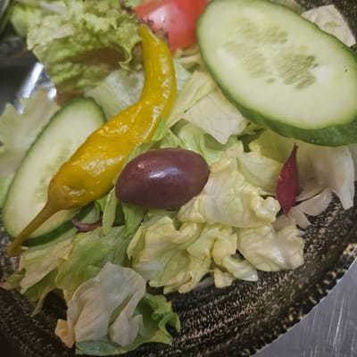 A close-up shot depicts a salad in a bowl. The salad contains lettuce, cucumber slices, a yellow pepper, an olive, and a small piece of tomato. The bowl is in focus and positioned in a well-lit environment. The image does not suggest a specific location in Fehmarn, Germany, nor does it include people or any explicit actions or interactions.