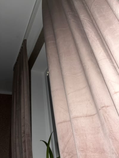 The image shows a close-up view of the interior of a room, likely a bedroom or living room, with a focus on the window area. Two sets of curtains are visible, one a darker shade of brown on the left and the other a lighter, dusty rose or taupe color on the right. The curtains appear to be made of a soft, possibly velvet or velour material, and are drawn partially open, revealing a sliver of a white window frame and a light blue wall beyond.

The lighting suggests it might be evening or a dimly lit interior. A green plant with long, slender leaves is partially visible in the lower right quadrant of the image, its leaves reaching upwards. The ceiling and a piece of trim are visible in the upper left of the frame. The overall impression is one of a domestic interior, possibly in Ponebel, Ukraine, suggesting a common household setting. There are no people or animals present in the image.