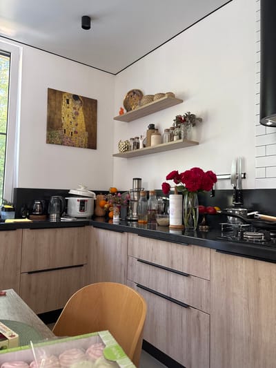 This is a medium shot of a modern kitchen with light wood cabinets and black countertops. The kitchen is located in Vardane-Verino, Russia. On the left side of the frame, a large window allows natural light to enter. Above the countertop, there are two wooden shelves displaying various items, including decorative baskets, jars, and a vase with flowers. A painting, "The Kiss" by Gustav Klimt, hangs on the wall to the left of the shelves. The countertop is filled with appliances and decorative items, such as a pressure cooker, electric kettles, bottles, and a vase of red roses. A bouquet of red roses in a glass vase is the central focus of the countertop arrangement. A knife rack with two knives is mounted on the backsplash. A black range hood with white tiled backsplash is visible on the far right. In the foreground, a wooden chair is partially visible next to a table with a box of sweets. The overall scene depicts a clean, well-organized, and aesthetically pleasing kitchen space.