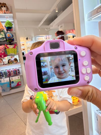 The image depicts a close-up of a person's hand holding a pink toy digital camera. The camera's screen displays a photograph of a young child smiling. In the background, a child wearing a white t-shirt is partially visible, holding a green and yellow toy. The environment appears to be a store, with shelves displaying various merchandise, including hats, clothing, and other toys. The overall lighting suggests an indoor retail setting. There is no specific indication of the city of Alboraia/Alboraya in Spain within the image.