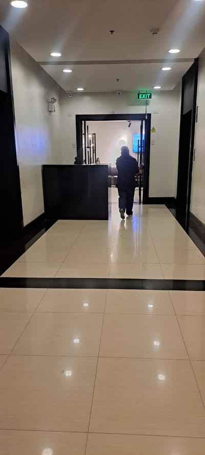 The image depicts a person walking down a brightly lit corridor, with polished beige floor tiles reflecting the overhead lights. The corridor features a black tiled stripe running vertically down the center, separating the tiled floor. On the left wall, there are several circular, recessed lights casting a white glow, and a green "EXIT" sign is prominently displayed, along with what appear to be security cameras and smoke detectors.

The person, dressed in dark clothing and a hood, is the sole subject in the visible frame. They are walking away from the camera, towards a doorway at the end of the corridor. Through the doorway, a brighter, possibly outdoor or more open space, can be seen, with a blueish glow emanating from within. A black counter or desk is positioned in front of the doorway.

The overall impression is one of a modern, well-maintained indoor environment, likely a commercial building or a similar public space. The lighting suggests it could be daytime or evening, as the artificial lights are the primary source of illumination. There are no overt indications of weather conditions. The scene is neutral, with no specific emotions conveyed beyond the solitary act of walking. The visible text is limited to the "EXIT" sign. The location context provided is Mandaluyong, Philippines.