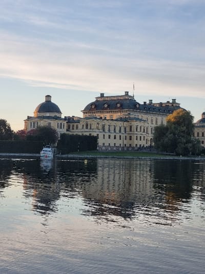 The image displays Drottningholm Palace, located on Lovön island in Ekerö kommun, Sweden, viewed across a body of water, likely Lake Mälaren. The palace is a large, multi-winged structure with a light yellow facade, dark gray curved roofs, and prominent domes on its peripheral sections. Numerous windows are visible across its multiple stories. The architectural details of the palace, along with the sky, are reflected in the subtly rippled water in the foreground. A white motorboat, featuring blue and red accent stripes, is moored near the palace's shoreline on the left side of the frame, positioned in front of dark green trees and hedges that line the property. The scene is illuminated by soft, warm, natural light from a pale blue sky with scattered wispy white clouds. No individuals are visible.