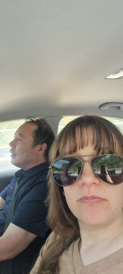 A woman with sunglasses is seated in a car with a man. The woman is wearing a tan-colored top and has long brown hair with bangs. The sunglasses are aviator style with a reflective lens. A man is sitting beside her in a navy-blue shirt and a seatbelt. The photo was taken inside the vehicle with a view to the outside. Based on the surroundings, the location is Bongpyeong-myeon, South Korea.
