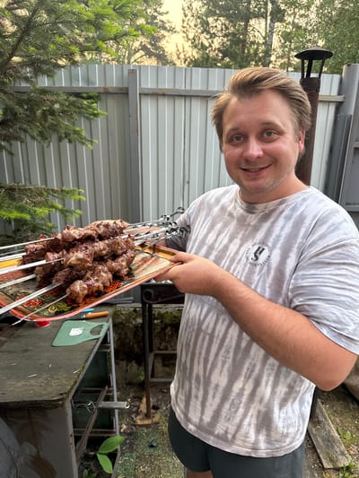 A Caucasian man with light brown hair and blue eyes, wearing a grey tie-dye t-shirt with a "High School Sports Club" logo and dark shorts, is holding a tray of meat skewers. He is smiling at the camera. The background shows a grey corrugated metal fence, green foliage from a coniferous tree and other trees, and a metal chimney. The man appears to be outdoors in a backyard or outdoor cooking area, possibly in a rural setting, like the village of Ust-Mana, Russia. There is a wooden table with a green cutting board and other objects on it in the foreground.