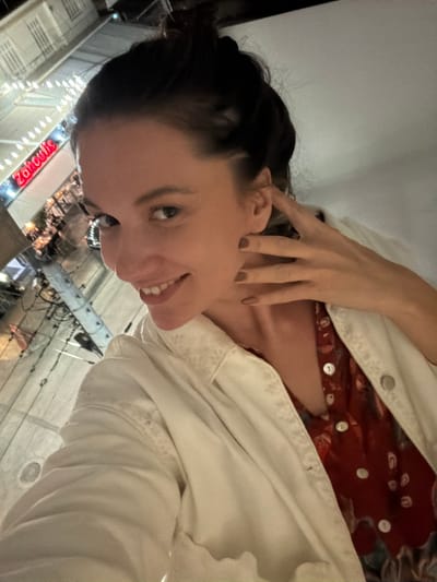 A selfie depicts a fair-skinned woman with dark hair pulled back, smiling slightly. She is wearing a red top with white polka dots, layered under a white jacket. Her left hand is near her face. The background features a street scene at night. A sign with the name "Zahoulis" is visible, identifying the location as Glifada, Greece. Other details include street lights and building facades. The perspective is angled upwards.
