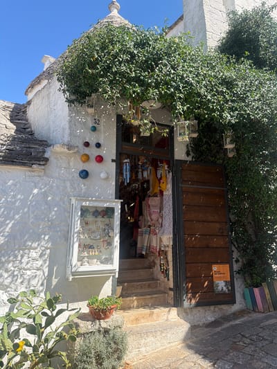 The image depicts a sun-drenched scene in Alberobello, Italy, focusing on the entrance of a shop nestled within a whitewashed, conical-roofed trullo. Lush green foliage cascades over the entrance, partially obscuring decorative lanterns hanging above. The trullo's rough-hewn stone roof and textured white walls are characteristic of the region's unique architecture.

On the left wall of the trullo, several colorful, spherical ceramic decorations are mounted, along with the number "32." Below these, a small glass-fronted cabinet displays an assortment of trinkets, including miniature houses, ceramic figures, and what appear to be jewelry items. In front of the cabinet, a prickly pear cactus with a yellow bloom stands next to a terracotta pot overflowing with green plants.

The shop entrance features a set of stone steps leading up to an open doorway, revealing a display of textiles and wind chimes within. To the right of the entrance is a large, rustic wooden door. A small orange sign is affixed to the door, bearing text in Italian that includes "Museo del Territorio ALBEROBELLO," suggesting the shop might be associated with a local museum or heritage site. A row of vibrantly colored wooden planks leans against the wall to the far right of the door. The ground in front of the trullo is paved with irregular stone, illuminated by dappled sunlight filtering through the leaves. The sky above is a clear, bright blue. No people are visible in the image.