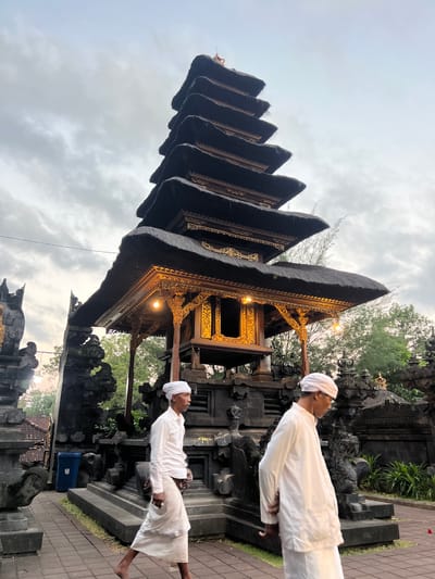 In Dawan, Indonesia, the image captures a multi-tiered pagoda structure with dark, thatched roofing and ornate golden accents. The pagoda is flanked by dark stone carvings and intricate stonework. Two men, dressed in traditional white attire consisting of long-sleeved shirts and sarongs, walk in the foreground. The man on the left is seen from the side, while the man on the right is also shown in profile. The sky above is overcast, with diffused light suggesting it is either early morning or late evening. The overall setting appears to be a temple or cultural site, with lush greenery visible in the background.