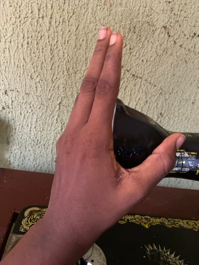 A hand firmly grasps a black bottle with a shiny label. The index and middle fingers are extended, creating a distinct separation from the other fingers. The hand rests on a textured, light-colored surface, with a portion of a dark-colored sewing machine featuring ornamental details visible on the right side. The composition is well-lit, emphasizing the contrast between the dark bottle and the lighter hand.