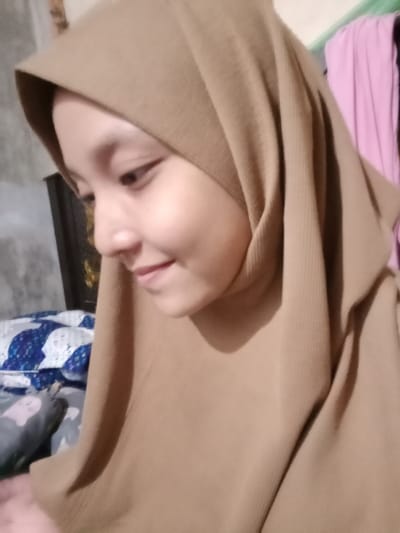 An indoor photograph depicts a young female individual in Lam Ujong, Indonesia. She is shown in a three-quarter profile, looking downwards and to the left, displaying a subtle smile. She wears a light brown, ribbed hijab covering her head and draping over her upper torso. Her right hand is partially visible in the lower left corner. The background to the left features a blue and white patterned pillow on a bed-like surface, with a grey object below it. A dark, vertical furniture piece is present behind her head. On the right side of the background, blurred pink and light purple fabrics hang vertically.
