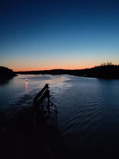 The image depicts a waterway at dusk, likely a river or a wide canal, in Badozhskoi Pogost, Russia. The foreground shows the dark silhouette of what appears to be the deck and crane of a vessel. The water is dark and rippled, reflecting faint light. Red and green navigation lights are visible on the water's surface, marking the channel. On either side of the waterway, the treeline is silhouetted against the horizon. The sky transitions from a deep blue at the top to orange and pink hues near the horizon, indicating either sunset or sunrise. There are no people visible in the scene. The focus is on the machinery of the vessel and the landscape.