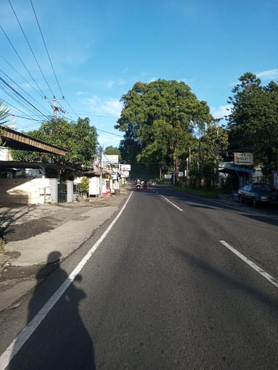 The image captures an outdoor street scene in Khouathineung, Laos, under a clear blue sky with sparse clouds, indicating bright daylight. The perspective is low, looking down an asphalt road that features a solid white line on the left edge and a dashed white line further ahead. Long, distinct shadows of at least two figures, possibly an adult and a child or a person on a small vehicle, are cast onto the concrete sidewalk and the left lane of the road in the foreground.
On the left side of the road, a series of low-rise buildings or stalls with various roofs and awnings line a concrete sidewalk, interspersed with green trees and utility poles supporting a dense network of overhead electrical wires. Farther along this side, additional commercial structures are visible. On the right side of the road, a dark-colored sedan is parked off the pavement, partially sheltered by a blue tarp or awning structure. This side is also bordered by lush green trees and foliage, with more utility poles. A white sign bearing black text, partially obscured by greenery, reads "SHOLAT ?" with an accompanying arrow pointing left. In the mid-distance, several motorcycles and at least one car are observed traveling away from the camera along the road. No specific interactions between individuals are discernible.