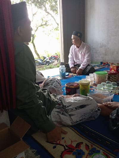 The image captures an indoor scene in Bulukandang, Indonesia, featuring two adult males seated on patterned floor mats. An open doorway in the background reveals an outdoor environment. The individual in the foreground, positioned to the left, wears a dark olive-green long-sleeved shirt and a dark peci, with a white patterned sarong covering his lower body. His right hand rests on a beige rug with red and blue floral motifs. The second individual, an older male, is seated cross-legged in the background to the right, wearing a light pink long-sleeved shirt and a black peci, looking towards the outdoor area. Between the two men, various items are arranged on the floor mats: a blue jar, a clear rectangular container filled with light brown granular material, a green lidded container, a dark cylindrical container, a yellow lidded container, and a stack of clear small glasses. A partial view of a cardboard box is visible in the lower-left corner. Outside the doorway, two motorcycles, one white/grey and one red, are parked on unpaved ground amidst green foliage. A red curtain-like fabric element is visible on the far left edge of the frame.