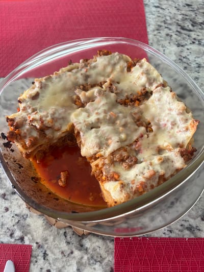The image shows a glass baking dish containing lasagna on a countertop. The lasagna appears to have layers of pasta, meat sauce, and melted cheese. A pool of tomato-based sauce surrounds the lasagna in the dish. The countertop is a light gray speckled surface. Part of a red woven placemat is visible on either side of the dish. There is no evidence to suggest this image was taken in Barrio de Nuevo León, Mexico. No people, actions, or interactions are visible.