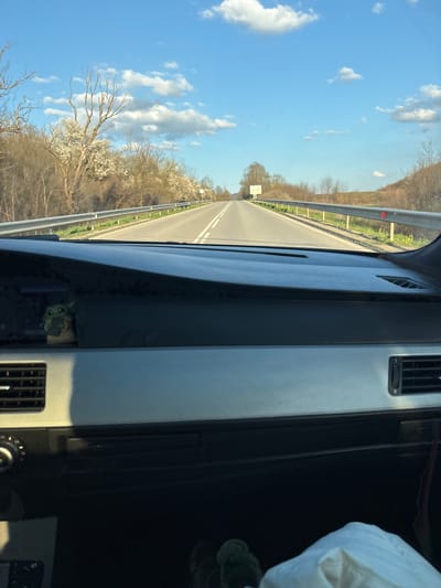 A view from inside a vehicle traveling on a road. The road is divided by a double white line. Beyond the road is a guardrail, trees, and a blue sky with white clouds. Inside the vehicle, the dashboard and air vents are visible. A figure resembling "Baby Yoda" is placed in a storage compartment. A white bag is partially visible in the foreground. The location is undetermined.