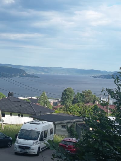 The image depicts a scenic view from a residential area overlooking a wide body of water, likely a fjord or a large lake, in Drammen, Norway. In the foreground, several houses with dark tiled roofs are visible, nestled amongst green trees. A white camper van is parked on a sloped driveway in front of a light-colored building, with a dark-colored car parked to its left. A red car is parked further down the driveway, partially obscured by foliage. The background features rolling hills and mountains that recede into the distance, meeting a hazy sky with scattered clouds. Utility lines are strung across the upper portion of the scene, dividing the sky from the landscape. The overall impression is one of a quiet, suburban setting with a beautiful natural backdrop.