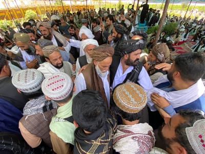 The image shows a large gathering of men in a crowded outdoor space. The location is in Zaker-E Sharif, Afghanistan. Many of the men are wearing traditional Afghan clothing, including turbans, long shirts, and loose trousers. There are several men in white long shirts and white turbans with intricate designs. The scene appears to be a religious gathering, with many of the men engaged in conversation and prayer. Some men are shaking hands and hugging, while others are sitting on the ground. The men are arranged in a large circle, and their faces are visible, and many of the men are looking at the camera. The ground is covered in dirt, and there is a large tent in the background. The scene is peaceful and serene, with a sense of community and religious devotion.