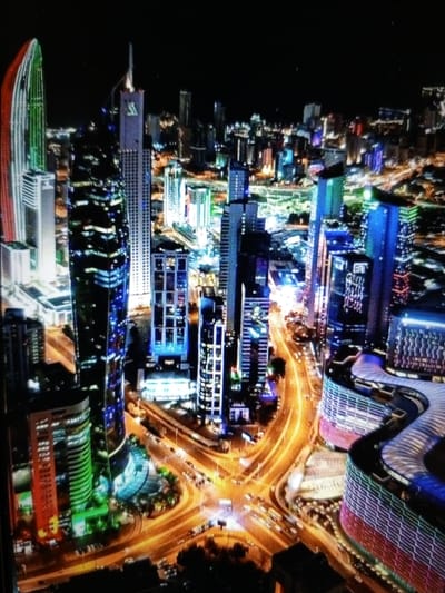 This is a nighttime aerial view of a modern cityscape, likely in a large urban center. The image is dominated by numerous skyscrapers of varying heights and architectural designs, illuminated by a multitude of lights. Many buildings feature decorative lighting, with some displaying vibrant colors like green, blue, and red. The roads below are visible, illuminated by streetlights and the headlights and taillights of moving vehicles, creating streaks of light that suggest the flow of traffic.
In the foreground and middle ground, several distinct buildings are prominent. To the left, a tall, curved skyscraper has a facade that appears to be a gradient of red and green lights. Adjacent to it is a slender, dark skyscraper with horizontal bands of light. Further into the scene, a cluster of buildings presents a mix of sharp geometric shapes and some with softer, curved exteriors. One building in the mid-right section of the image has a notable curved facade, patterned with horizontal bands of purple and pinkish-red lights. Another nearby structure features a rounded, multi-tiered design with a light blue illumination. The overall impression is one of a bustling and technologically advanced urban environment at night. The specific location in Golbembal, Nigeria, cannot be determined from the visual information provided in the image.