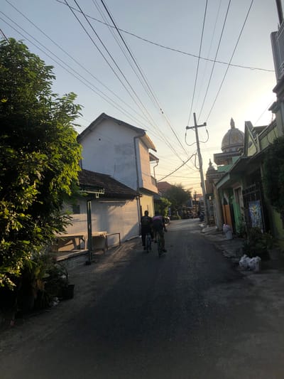 In the city of Putat, Indonesia, a paved road winds through a residential area under a clear sky at either dawn or dusk. Two individuals are cycling away from the camera on the road. On the left side of the road, a lush green tree with dense foliage partially obscures a white, two-story house with a tiled roof. In front of this house, there are two wooden tables with benches.

On the right side of the road, several buildings are visible. One building features a distinctive domed roof, characteristic of a mosque or a religious structure. This building is adorned with intricate details and appears to be constructed from stone and possibly painted. Adjacent to it are other houses with green facades and tiled roofs. Power lines crisscross the sky above the street, supported by utility poles. The lighting suggests either early morning or late afternoon, casting long shadows. There are potted plants and what appear to be bags of trash on the side of the road.