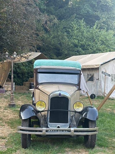 A vintage, two-tone beige and brown truck with a green tarp draped over its roof is parked on a grassy area. The truck has a chrome grille, two yellow headlights, and a chrome bumper. It is facing forward, with its front wheels on the grass. In the background, there are several canvas tents, hay bales, and trees. A vintage lamppost with three lights stands to the left of the truck. A wooden sign with arrow pointing right, with the word "TUTTI LE", is attached to a wooden pole to the right of the truck. The scene appears to be set up for an outdoor event or festival. The location is Saint-Vaast-du-Val, France. There are no people visible in the image.