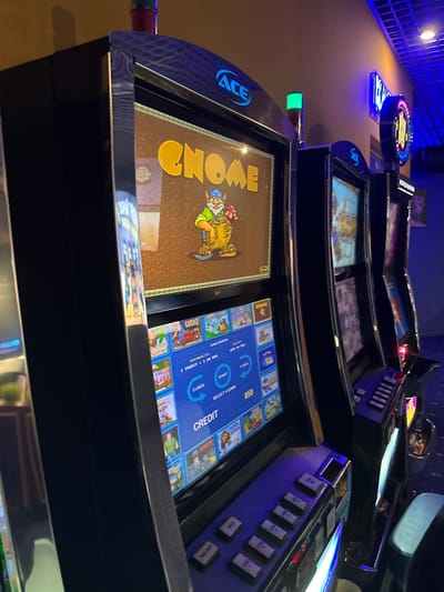 The image shows a row of slot machines in what appears to be a casino or gaming establishment. The machines are black with metallic trim and feature large screens displaying colorful graphics. The top screen of the leftmost machine shows a cartoon character, possibly a gnome, with the word "Gnome" written in large yellow letters. The lower screen displays game options, including "Garage," "Sweet Life," and "Rock Climber," along with details about credits and lines. The machines are lit by a combination of overhead lighting and the screens themselves, with blue and purple hues prominent. Some of the machines have illuminated signage at the top, including one with the "ACE" logo. The overall environment suggests a dimly lit, modern entertainment space. There are no people visible in the image. The city of Lotnoye, Russia, is not depicted or referenced in the image.