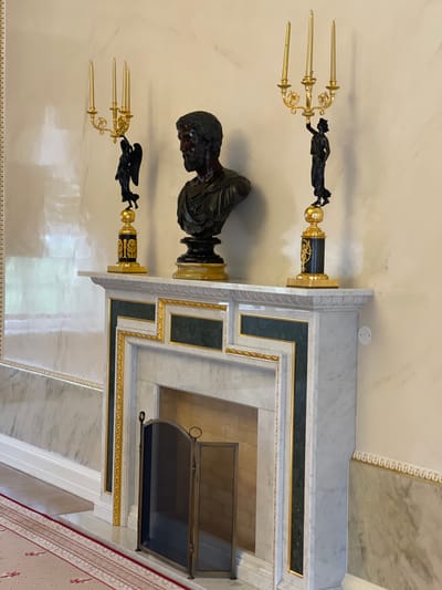 The image depicts an ornate marble fireplace in a well-lit room. The fireplace mantel is adorned with two golden candelabras, each featuring three candles and a black statuette of a winged angel on the left and a robed female figure on the right. Centered on the mantelpiece is a dark bronze bust of a man with a beard, wearing a draped garment. The fireplace opening is covered by a dark metal screen. The walls are a pale, textured color, with marble accents and decorative golden trim. A patterned carpet is visible in the foreground. This scene is located in the Grand Ballroom of Konstantinovsky Palace in Strelna, Russia.
