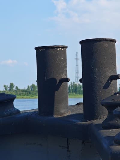 The image shows two black bollards on the deck of a vessel, with a body of water and a treeline in the background. The sky is blue with scattered clouds. A tall transmission tower is visible in the distance. The bollards are cylindrical with flared tops and have small metal handles protruding from their sides. The vessel's deck is also painted black and appears weathered. The overall scene suggests an industrial or maritime setting.